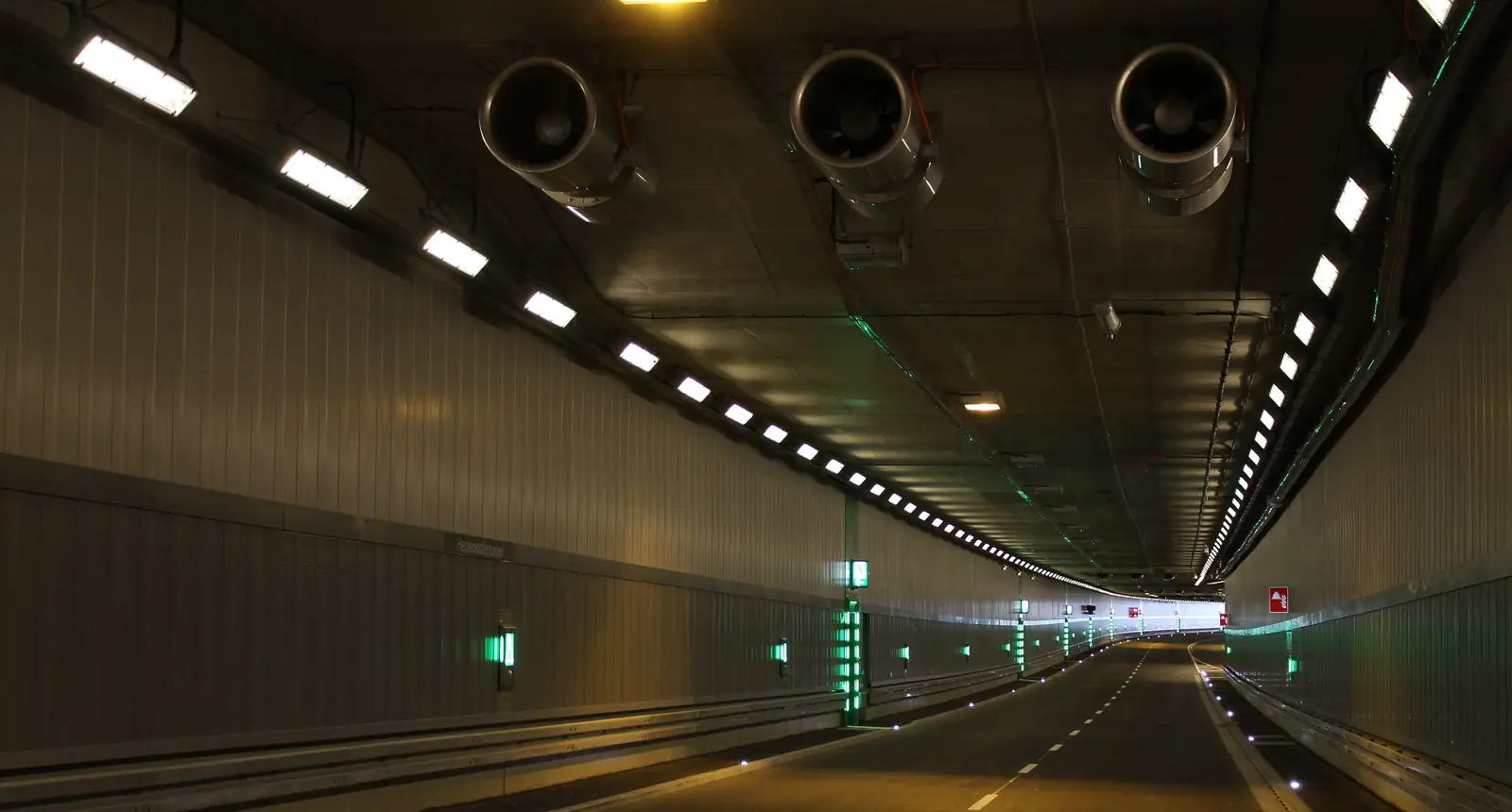 Interior de un túnel vial con ventiladores de chorro suspendidos de la bóveda y luminarias a lo largo del hastial.