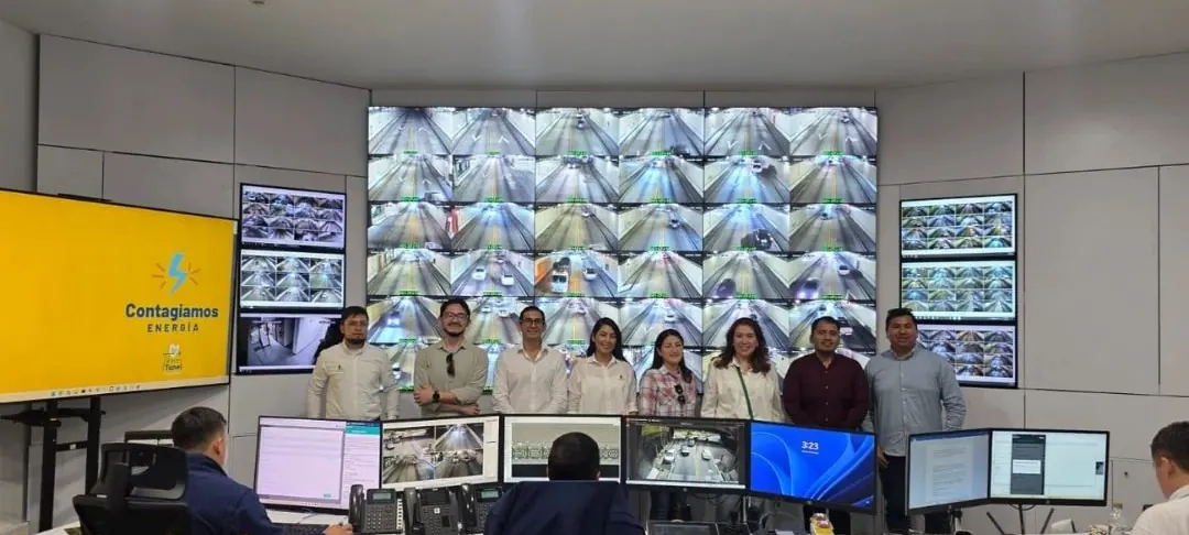 Sala de control de un túnel con un muro de video CCTV; visitantes de la delegación técnica de pie frente a los puestos de operación.